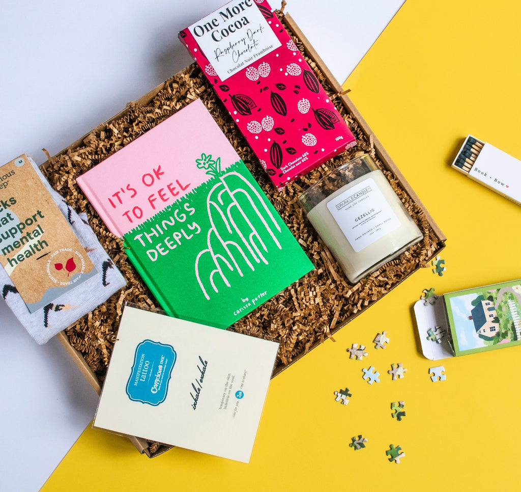 Image of a gift box with the book "It's Ok to Feel Things Deeply", a candle, a mini puzzle, an "inhale/exhale" temporary tattoo, a dark chocolate raspberry chocolate bar and socks that support mental mental health with people doing yoga on the socks. 